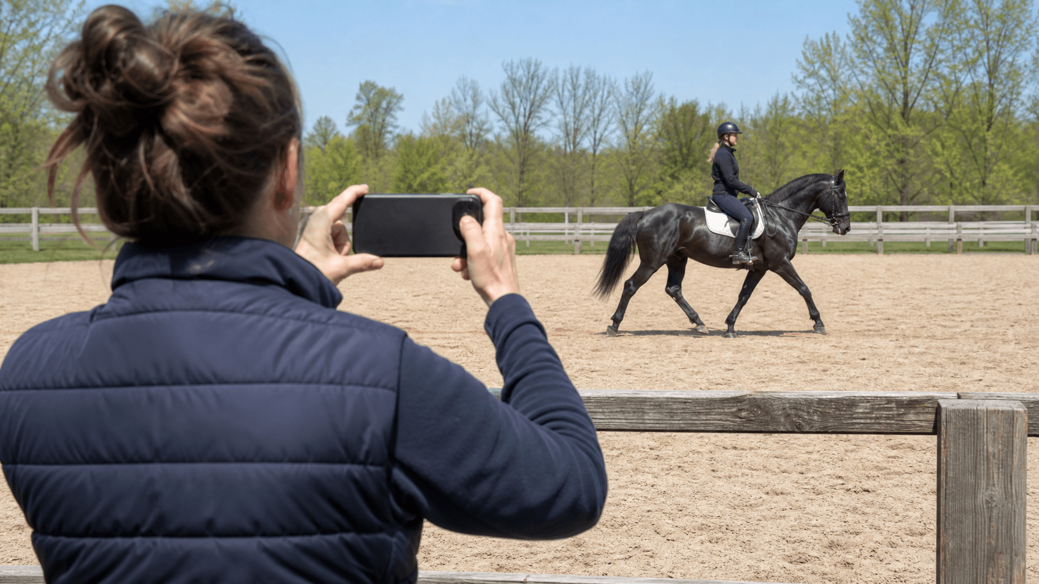A helper at the arena rail films a horse and rider with a phone held sideways in landscape orientation. The whole horse and rider are in frame and lit from the front.